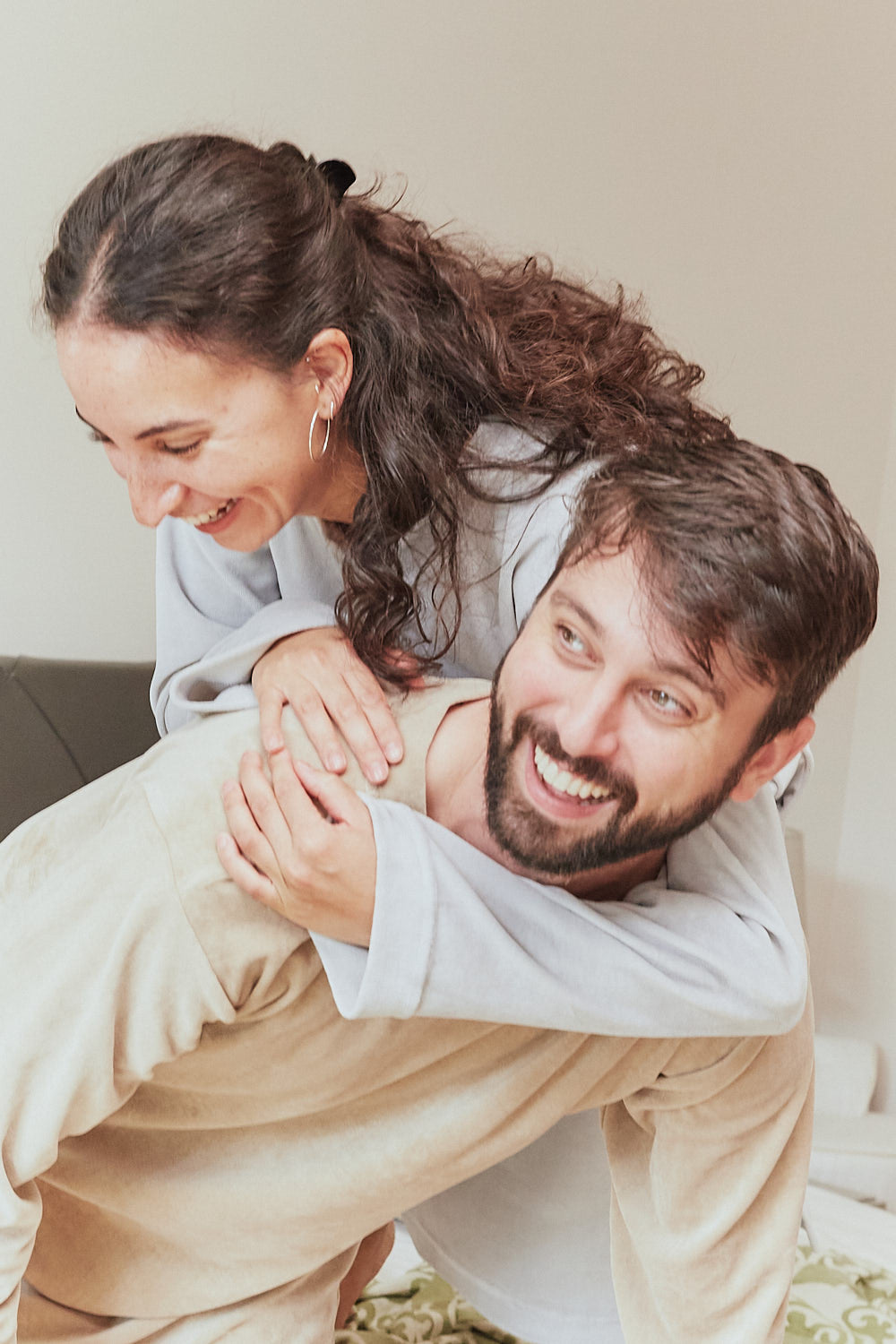 Lachendes Paar in Sonus Aeris Unisex Einteilern beim Spielen im Wohnzimmer cozy nachhaltige Loungewear fuer Sofa und Zuhause