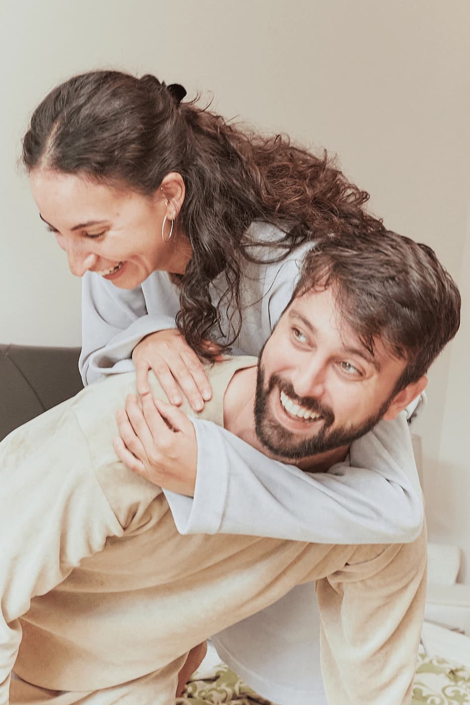 Lachendes Paar in Sonus Aeris Nicki-Velours Einteilern beim spielerischen Herumtragen im Wohnzimmer