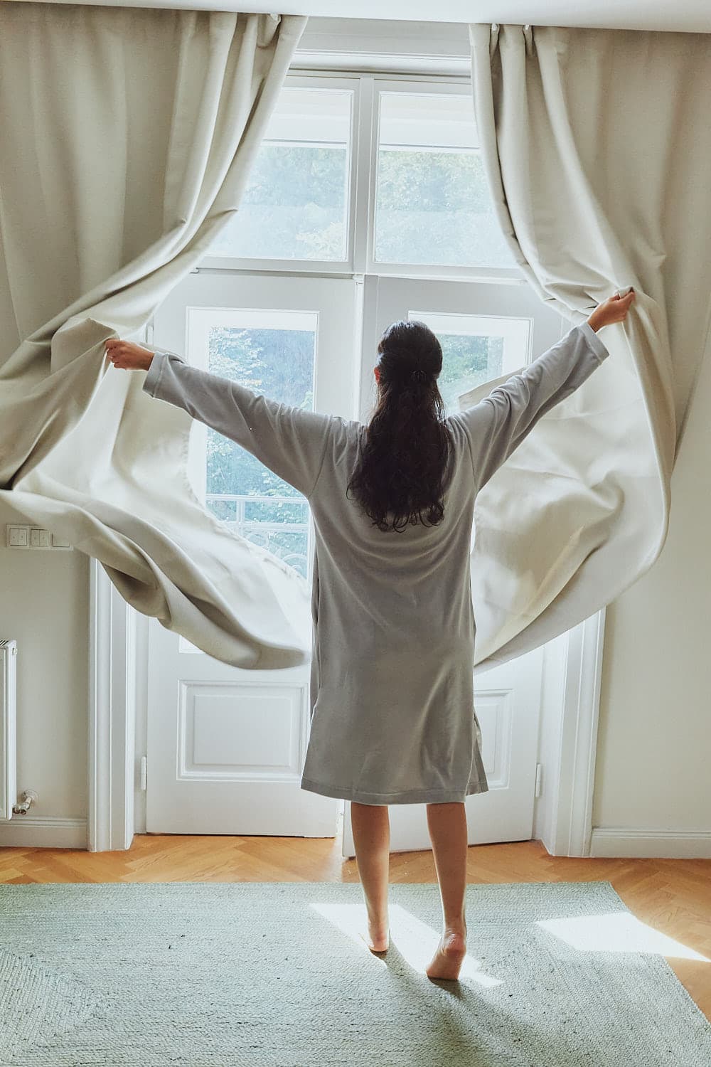 Person von hinten im Sonus Aeris Einteiler, steht am Fenster und zieht die Vorhänge auf.