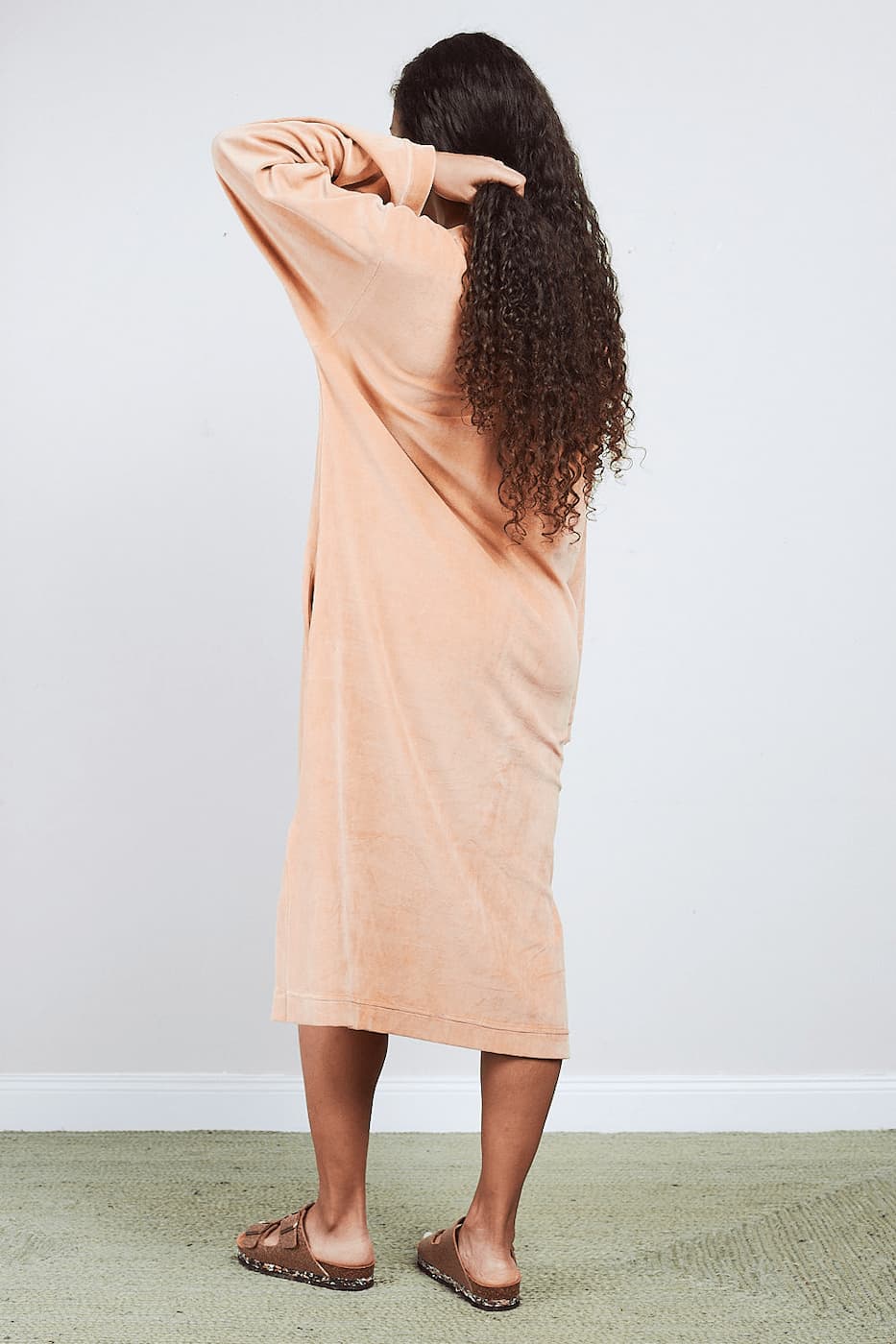 Rückansicht einer Frau im sandfarbenen Sonus Aeris Nicki-Velours Einteiler, die ihr lockiges Haar richtet