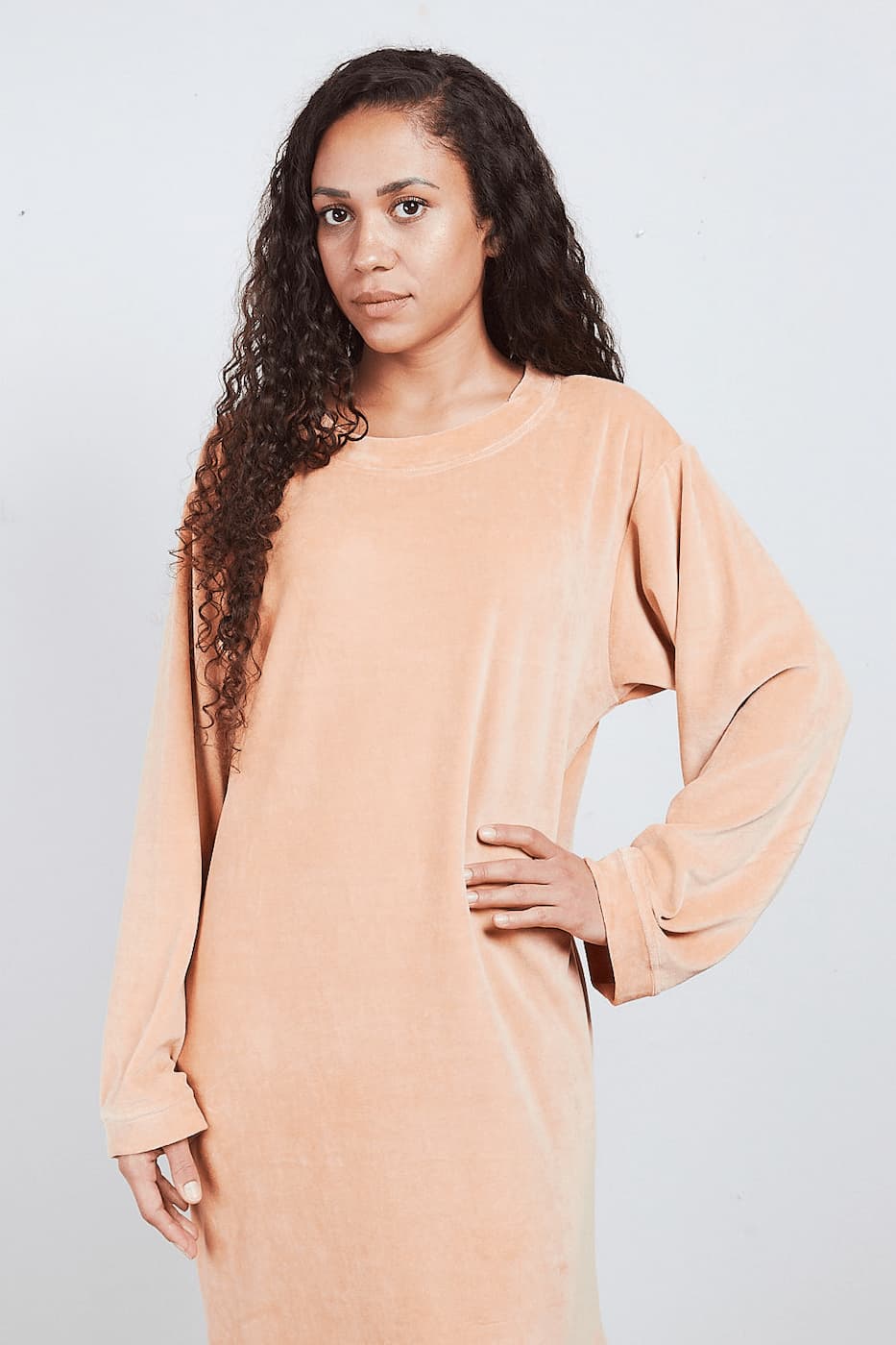 Frau im sandfarbenen Sonus Aeris Nicki-Velours Einteiler mit Hand in der Hüfte vor hellem Hintergrund