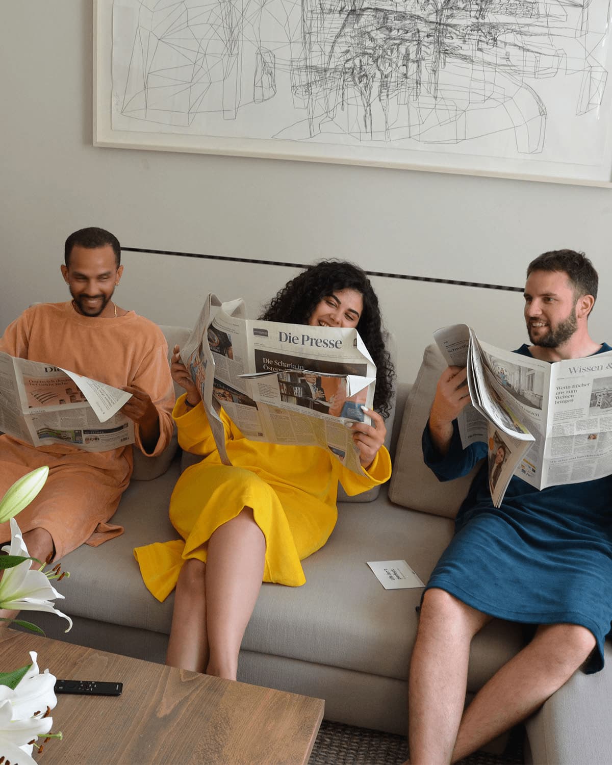 Three friends in Sonus Aeris one-pieces reading newspapers on the sofa